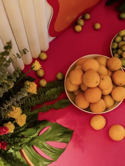 A top-down detail shot of the Mehendi decor, showing bowls of fresh oranges and limes scattered on a vibrant pink surface next to a lush floral arrangement.