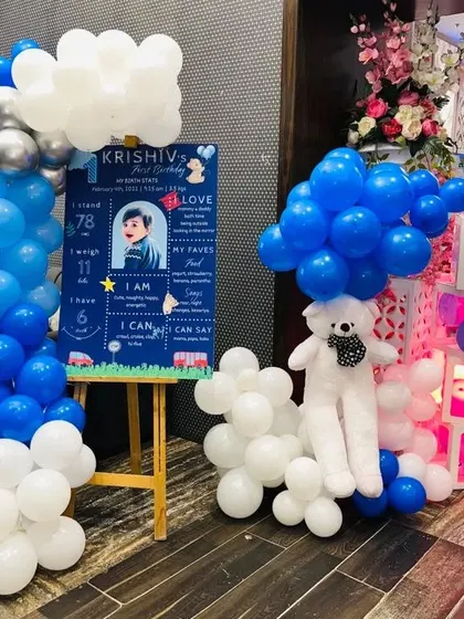 A personalized first birthday corner at home. The setup includes a milestone board, balloon arrangements, "BABY" blocks, and a cute white teddy bear.