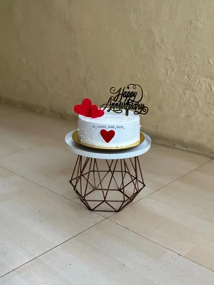 A simple and elegant anniversary cake with red hearts and a "Happy Anniversary" topper.
