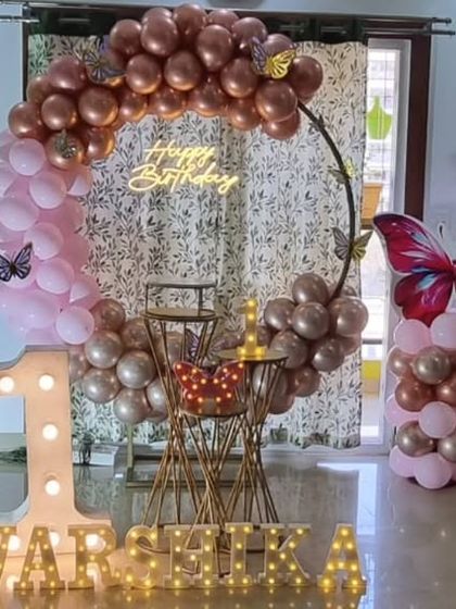 A chic butterfly-themed first birthday decoration for a home setting. The design uses a rose gold and pink balloon ring, a large marquee '1', and butterfly accents.