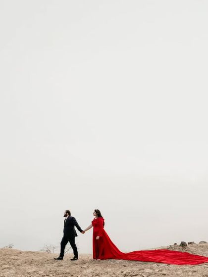 A minimalist composition from a destination pre-wedding shoot. The couple walks hand-in-hand along a ridge, with the long trail of the red dress creating a striking line in the landscape.