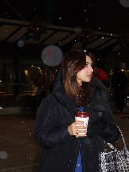 A candid shot in the falling snow at night, holding a coffee cup. This creates a cozy and magical winter moment.