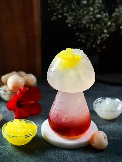 A creative lychee-flavored drink in a mushroom-shaped glass, showcasing layers of color and popping boba. The moody lighting and unique presentation make this a standout shot for a creative menu.