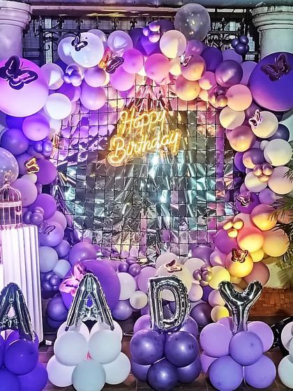 An outdoor birthday setup with a purple and white balloon arch against a silver fringe backdrop, perfect for an evening celebration.