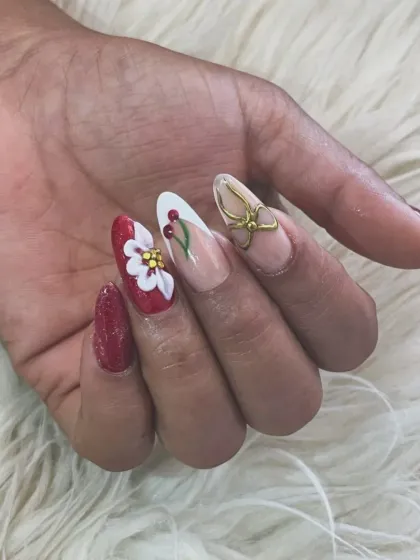 A playful and detailed set of 3D nail art. Featuring a mix of glossy red, a hand-sculpted white flower with a yellow center, delicate berry art, and a gold 3D bow accent on almond-shaped extensions.