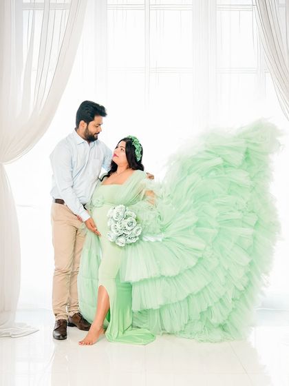 A sweet couple's portrait in our bright, window-lit studio space. The mom-to-be wears a voluminous mint green ruffled gown, creating a soft and romantic image.