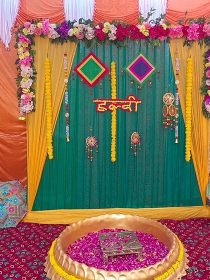 A traditional Haldi ceremony setup with a large brass urli filled with rose petals. The backdrop features green and yellow drapes, marigold garlands, and colorful hangings.