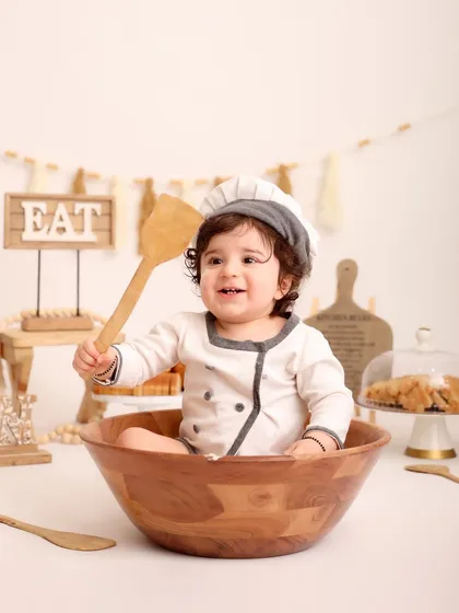 This little chef, sitting in a large wooden bowl and holding a spatula, smiles brightly for the camera. The setup is complete with rustic kitchen props, creating an adorable and memorable scene.