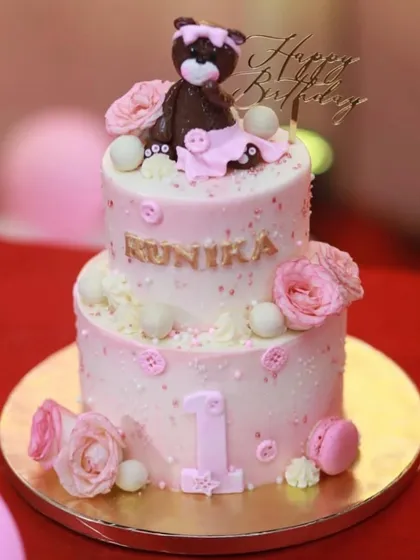 A beautiful two-tiered pink cake for a first birthday. This design features a chocolate teddy bear, fondant roses, macarons, and delicate button details.