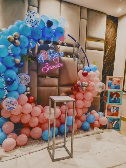 A baby shower setup with a pink and blue balloon ring, a neon sign, and "BABY" blocks, set against a modern padded wall.