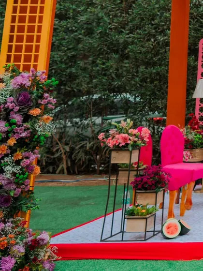 A side view of the Mehendi stage, showcasing the layers of colour from the orange structure, pink jaali, and lush, multi-coloured floral arrangements.