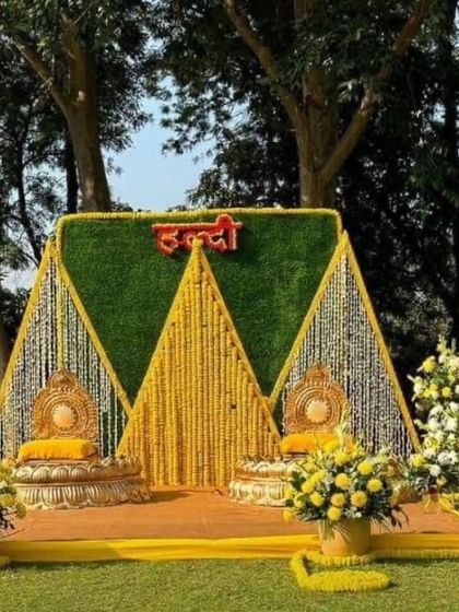 A unique triangular backdrop for an outdoor Haldi ceremony. The structure uses a green foliage panel and is decorated with strings of marigold and white flowers, with traditional seating in front.
