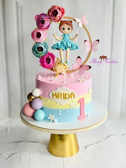 A dreamy pastel rainbow cake for Hrida's first birthday. The design features a doll on a floral swing, delicate butterflies, and soft colors for a truly whimsical celebration.