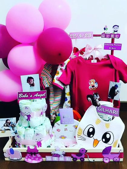 This personalized hamper for a baby girl features a diaper cake, multiple outfits, and custom photo cards. The vibrant pink theme and 'Gilhari' (squirrel) details make it a truly unique and personal welcome gift.