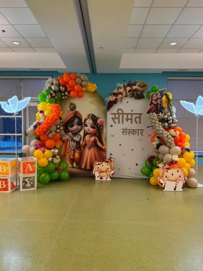 This Seemantha Sanskar setup uses a bright and cheerful color palette. The decor includes character cutouts, glowing butterfly lights, and a multi-colored balloon arch with orange, yellow, green, and silver balloons.