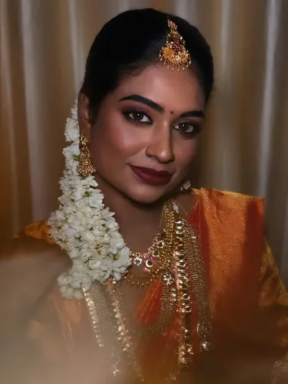 A close-up of a model with dusky skin, showcasing a traditional look with bold eyes and a deep red lip. The jasmine flowers in her hair complete the look.