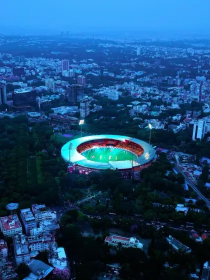An aerial view of the M. Chinnaswamy Stadium at dusk, the heart of cricket in Bengaluru. This shot captures the stadium nestled within the city's green canopy, lit up and ready for action, showcasing its iconic status.
