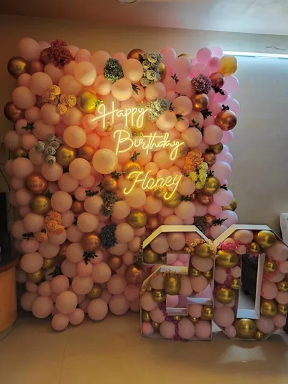 I can create a full floor-to-ceiling balloon wall for a stunning birthday surprise at home. This example uses a soft pink and gold color scheme, floral accents, and a personalized 'Happy Birthday Honey' neon sign.