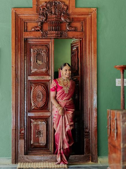 A bride in a vibrant pink saree, framed by an intricately carved antique door, a shot that is both regal and joyful.