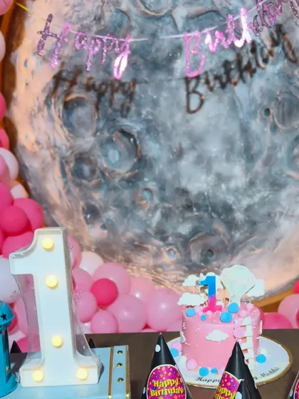 A close-up of the cake table at a moon-themed first birthday. The setup includes a themed cake, a light-up number '1', and custom party hats.