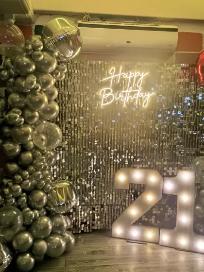 A stylish 21st birthday setup featuring a silver shimmer wall, a "Happy Birthday" neon sign, and a chrome silver balloon garland. The large light-up marquee numbers make a bold statement.