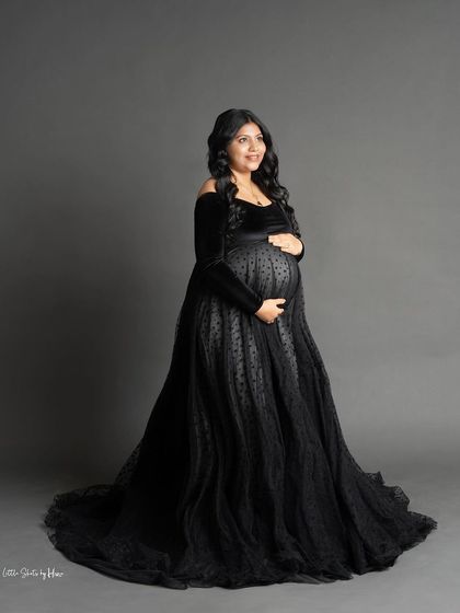 An elegant and moody portrait featuring a stunning black off-the-shoulder gown. The dress has a full, flowing skirt, and the mother-to-be looks off to the side with a gentle smile.