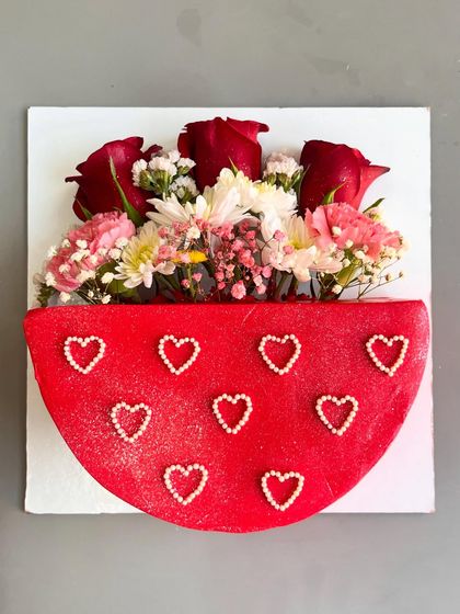 Elegance in every petal, sweetness in every bite. This unique semi-circular cake is designed to look like a bouquet holder, with a vibrant red base adorned with pearl hearts and an arrangement of fresh red and pink flowers on top.