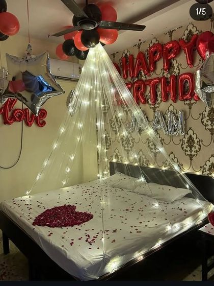 A romantic bed decoration with a canopy of fairy lights, a heart made of rose petals, and "Happy Birthday" foil balloons on the wall.