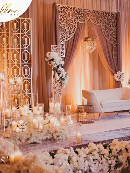 An all-white reception stage that exudes serenity and elegance. The decor features a wall of white flowers, candles, and intricate laser-cut panels for a modern yet timeless feel.