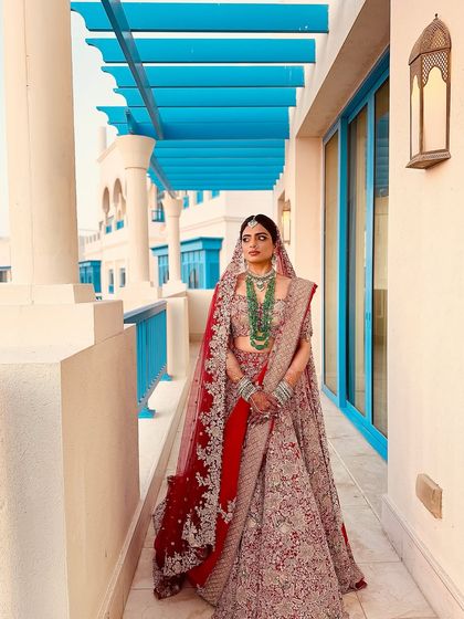 This bride looks absolutely regal against the stunning blue architecture in Doha. Her traditional red bridal lehenga is paired with makeup that highlights her features with a soft glow, perfect for a sunny destination wedding.