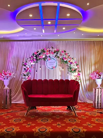 A wide shot of the complete stage decoration. The red velvet sofa and floral arch are flanked by matching flower arrangements on pedestals, creating a balanced and grand look for a formal celebration.