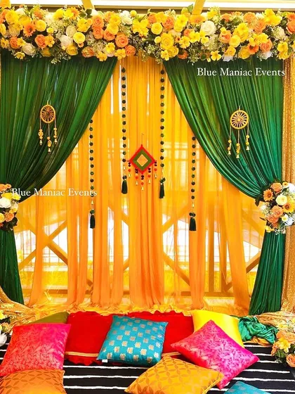 A colorful floor-seating Mehendi or Haldi setup. The backdrop of green and yellow drapes is complemented by bright, traditional cushions for a comfortable and festive arrangement.