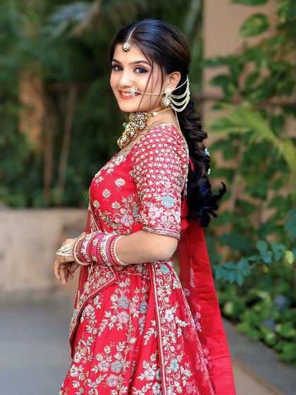 A side profile of a bride in a red lehenga, highlighting the detailed embroidery on the sleeves and the elegant pearl-drop earrings.