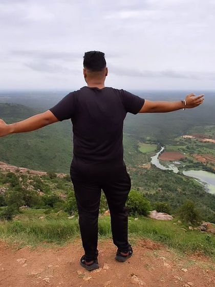 The feeling of freedom at the top of Makalidurga. A trekker spreads his arms wide, overlooking the winding river and green landscape below.