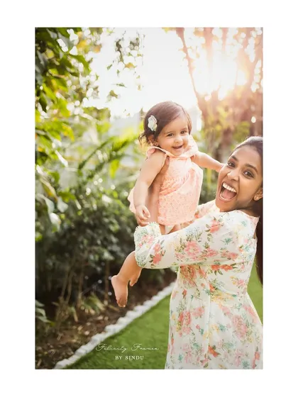 A mother joyfully lifts her laughing baby girl, both beaming with happiness. This golden hour outdoor shot is filled with warmth and pure, infectious joy.