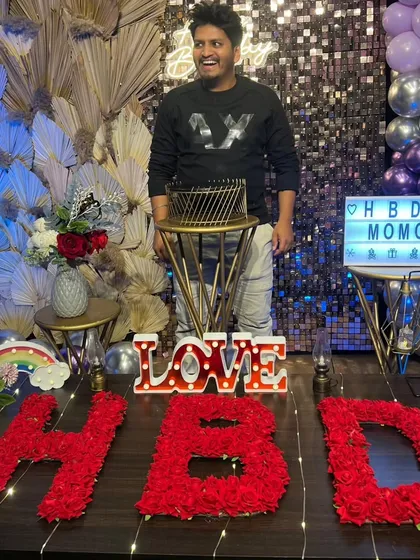 The birthday boy, "Momo," is all smiles as he stands with his cake. The custom lightbox, floral "HBD" letters, and sequin backdrop make for a picture-perfect birthday moment.