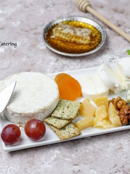 A pristine white platter showcasing a premium selection of cheeses including Brie, blue cheese, and goat cheese, served with crackers, dried fruit, and a side of honeycomb. This is a duplicate of image 12, presented here for its thematic fit.