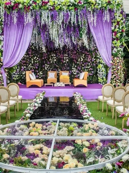 A truly surreal Jaimala ceremony setup featuring a transparent acrylic stage filled with flowers. The mandap is draped in shades of lavender and overflowing with wisteria and other purple blooms, creating a fairytale moment.