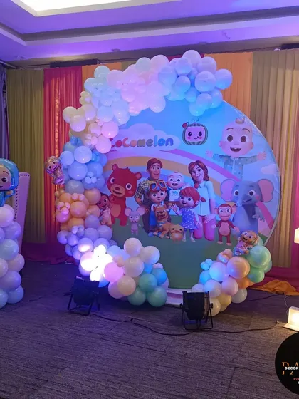 A Cocomelon-themed stage in a banquet hall. The large circular backdrop is framed by a pastel balloon arch and complemented by a light-up number "1" and character props.