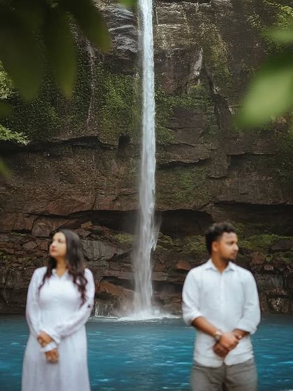 A formal, posed portrait set against the dramatic backdrop of a tall, slender waterfall. This image showcases a more contemplative and classic style of pre-wedding photography.