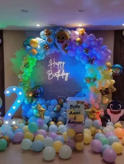 A vibrant Baby Shark themed birthday party. The decoration features a multi-colored balloon arch, a "Happy Birthday" neon sign, and character cutouts, creating an underwater feel.