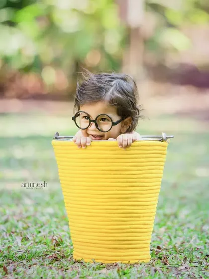 A fun outdoor shot of a toddler with glasses peeking out of a yellow bucket. Combining studio props with natural settings can create unique and playful images.