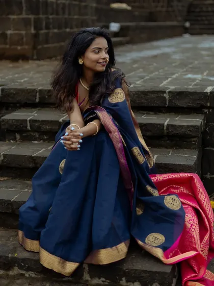 A full-length shot showcasing the beautiful drape and fall of a traditional blue and pink saree. The model is seated on stone steps, allowing the rich fabric and its border to be the main focus.