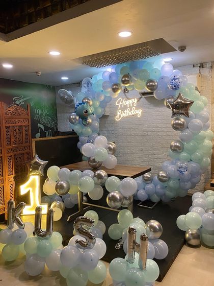 A dinosaur-themed first birthday party. The setup features a circular balloon arch in pastel blue and green, a light-up number '1', and a cute dinosaur foil balloon.