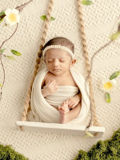 A serene and natural newborn portrait. The baby is swaddled in white and posed on a tiny swing, surrounded by moss and delicate white flowers for an ethereal feel.