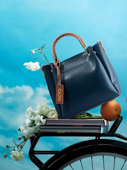 A creative and whimsical shot of a navy blue handbag resting on a bicycle, adorned with fresh flowers, against a bright blue sky.