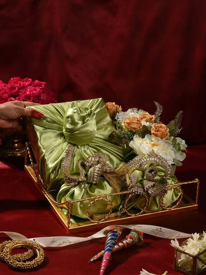 An elegant Teej hamper featuring gifts wrapped in beautiful green satin furoshiki-style cloth, arranged on a golden tray with flowers and traditional bangles.