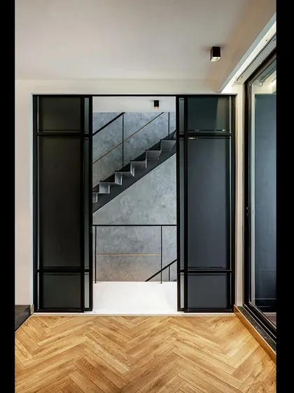 A view through a doorway in 'SOL' reveals the intricate layering of spaces. The herringbone wood floor of the room contrasts with the starker materials of the hallway and staircase, creating a clear threshold between different functional zones.