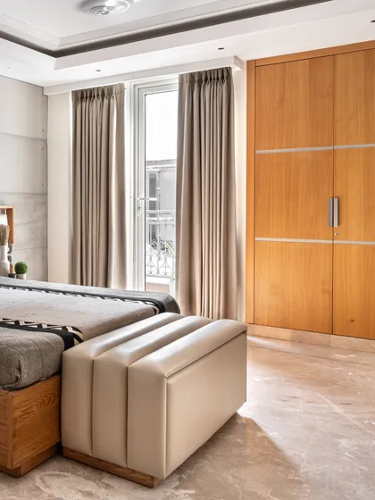 The room is filled with natural light from the large window, which is framed by simple, elegant curtains. The wooden wardrobe provides ample storage while maintaining the room's clean and minimal aesthetic.