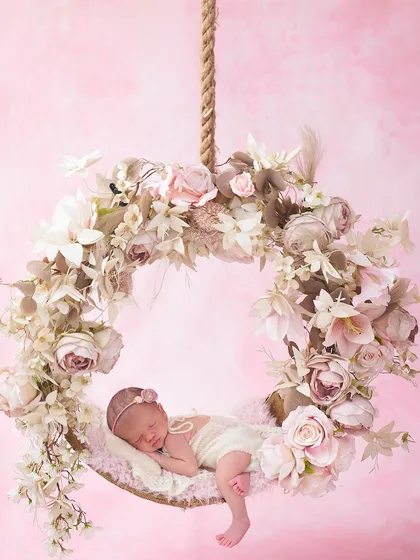 A baby girl in a white outfit sleeps on a hanging floral wreath made of muted pink and cream flowers against a soft pink background.